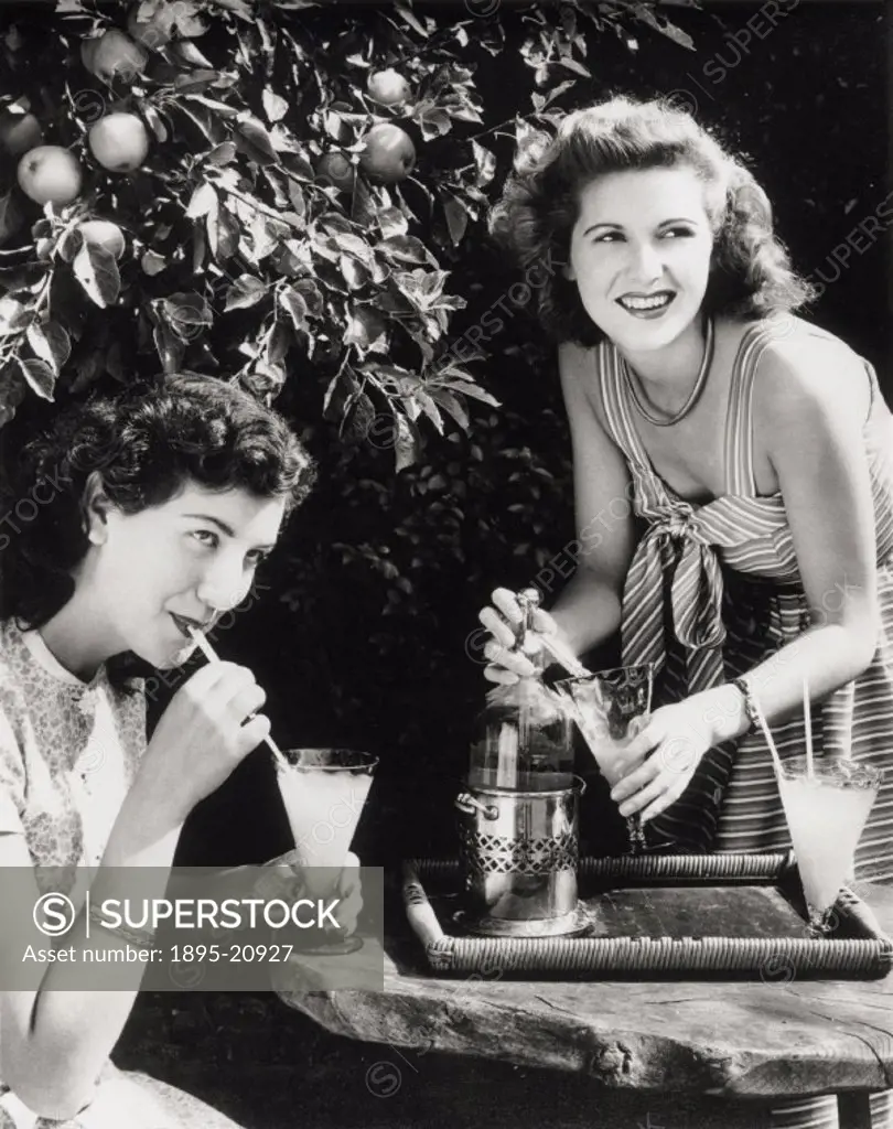 One of the women is drinking through a straw, while the other uses a soda syphon to fill her glass. An apple tree is behind them. One of a series of p...