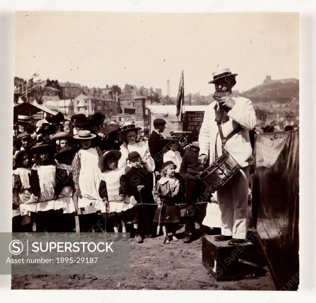 Photograph by Frank Meadow Sutcliffe (1853-1941) of a beach entertainer surrounded by a large audience of children. The man stands on a box playing a ...