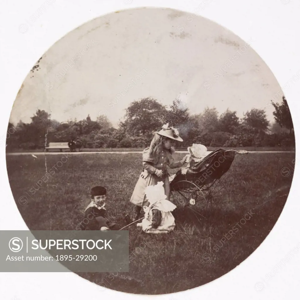 A Kodak circular snapshot photograph of three young children playing in a park, together with a baby in a pram, taken by an unknown photographer in ab...
