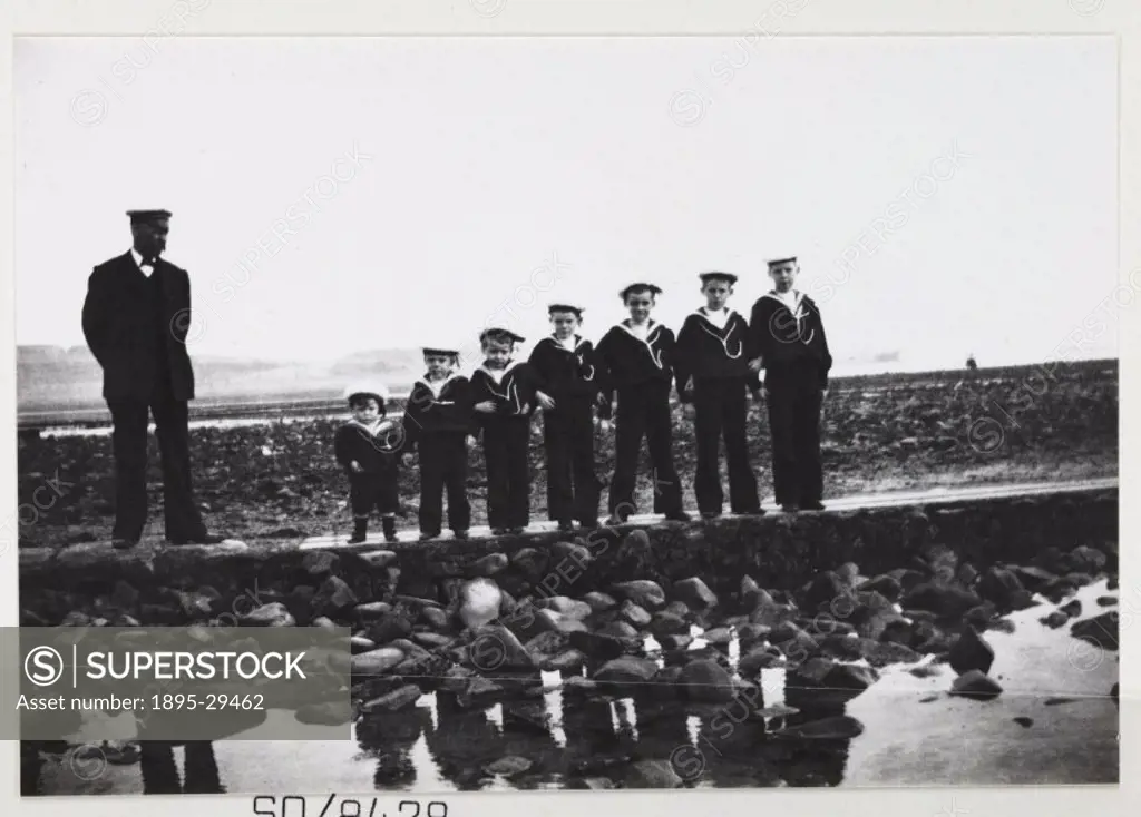 A snapshot photograph of a man and group of boys on the beach, taken by an unknown photographer in about 1900.  Seven young Sea Cadets line up in heig...