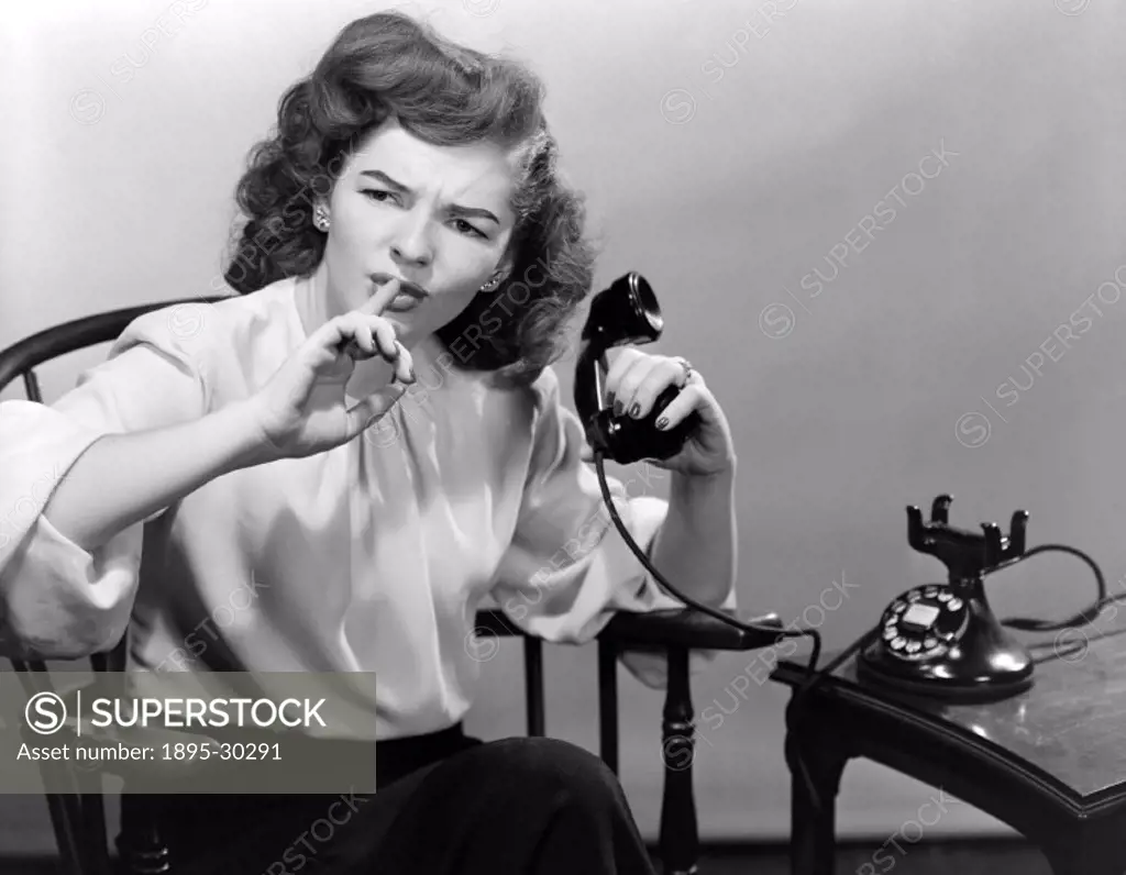 A photograph of a woman using the telephone, taken by Photographic Advertising Limited in about 1950.  The woman is making a hushing motion, holding h...