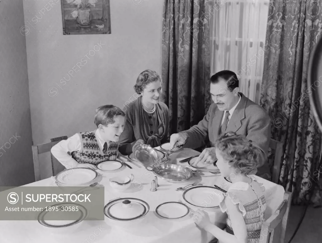 A photograph of a family about to eat dinner, taken by Photographic Advertising Limited in 1953.  Mother lifts the lid on the roasting tin so father c...