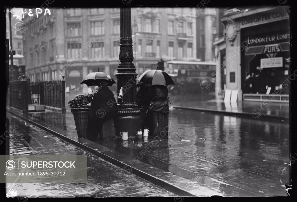 A photograph of flower sellers huddled beneath umbrellas on a wet London street, taken by James Jarche (1891-1965) for the Daily Herald newspaper on 2...