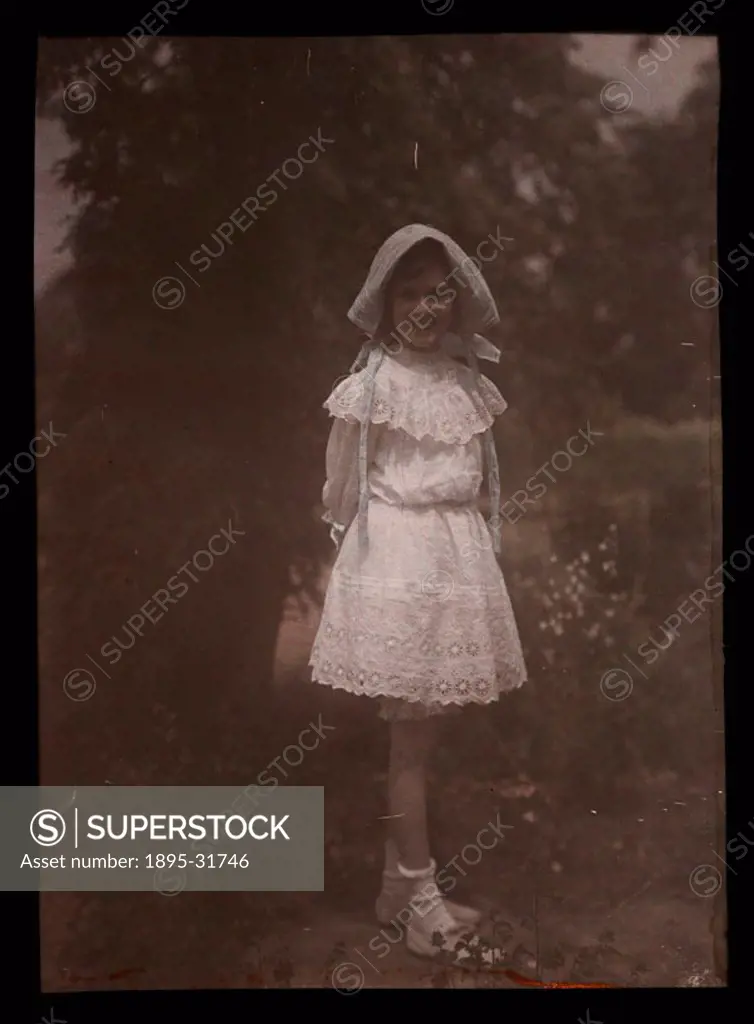 An autochrome of a smiling young girl, daughter of the photographer, wearing a bonnet and lace dress, standing in a garden, taken by Etheldreda Janet ...
