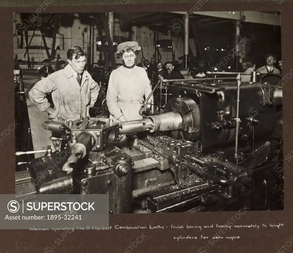 A photograph taken by an unknown photographer during 1915-1918 of a woman operating a machine lathe in a British munitions factory.  A male supervisor...