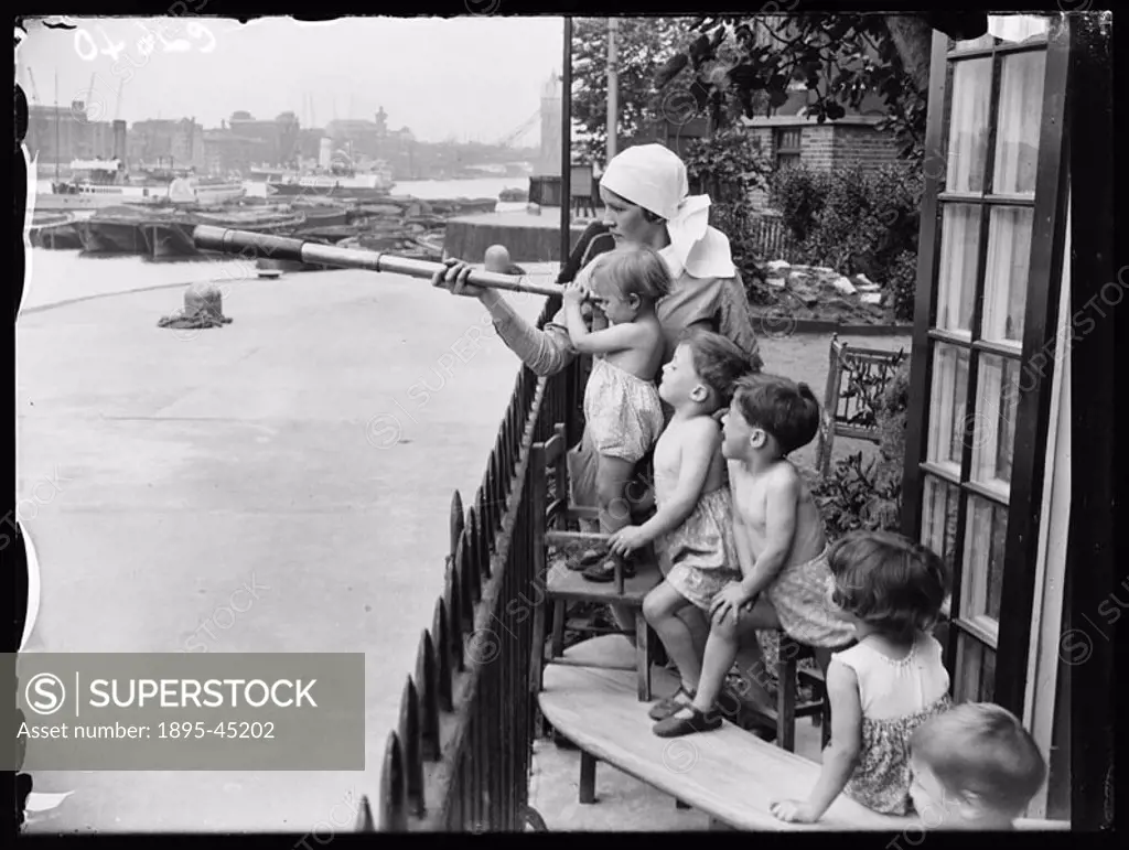 A nursery nurse holds a telescope while children look out at the River Thames. Tower Bridge can just be seen in the distance.