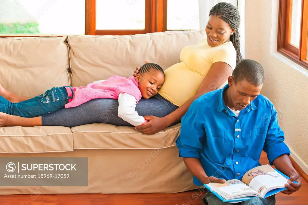 Girl lying on pregnant mothers stomach while her father is reading to them, Johannesburg, Gauteng Province, South Africa