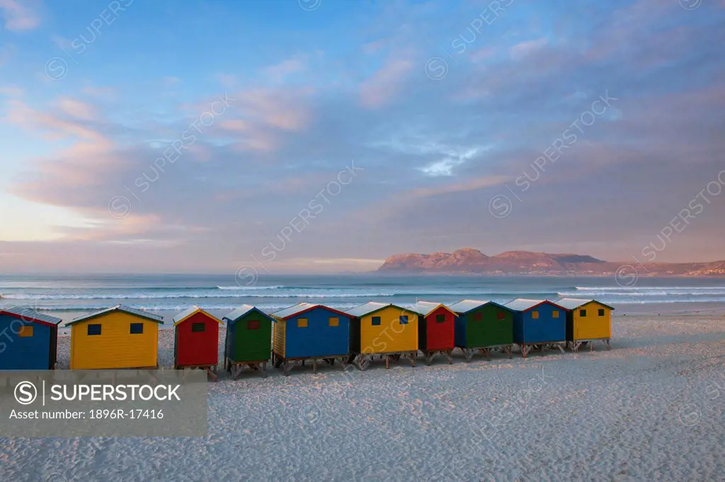 Colourful huts on white sands with the pink suNoise reflecting off the clouds and mountains in the distance. Muizenberg beach, Western Cape, South Afr...