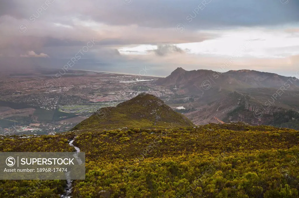 Footpath leading down mountain through thick green fynbos vegetation, suburbs of Constantia, Tokai and Muizenberg visible below with storm clouds and ...