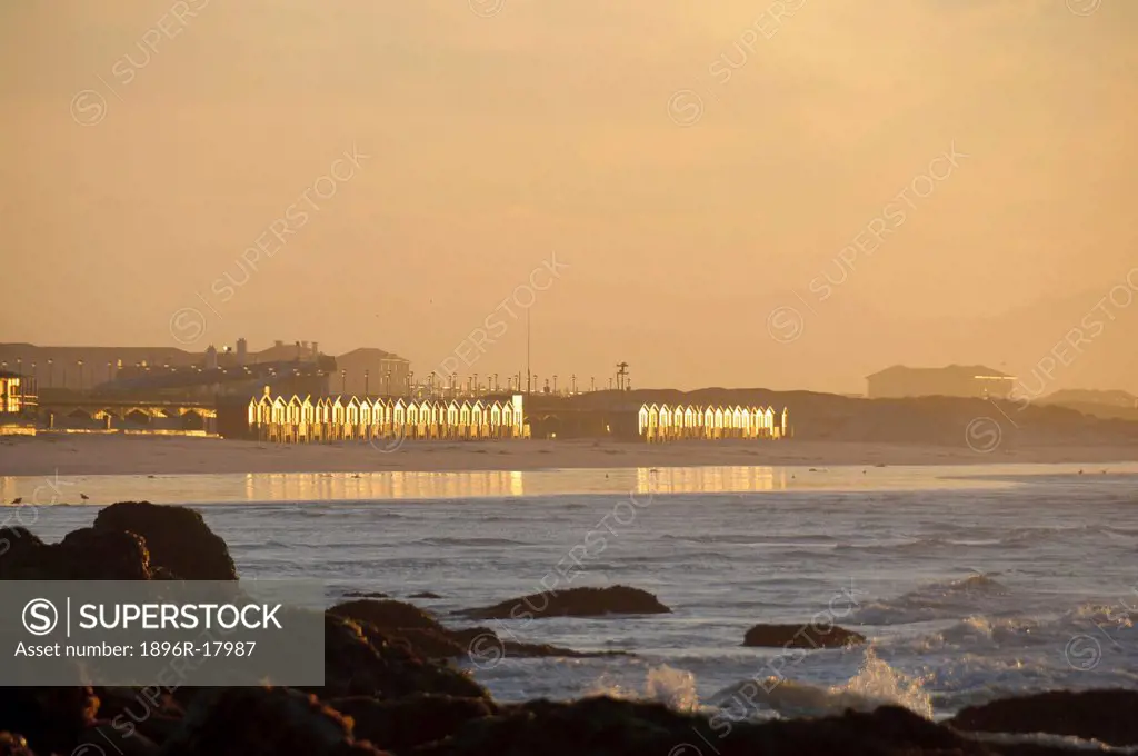 The iconic bath huts on Muizenberg beach reflect early morning suNoise, water slide in distance, rocks and sea in foreground, Muizenberg, Cape Town, W...