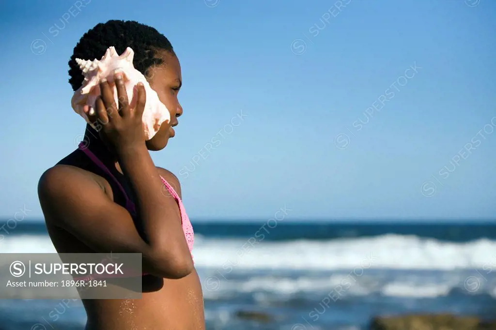 Young girl holds giant shell against her ear as she looks out at the Indian Ocean, KwaZulu Natal Province, South Africa
