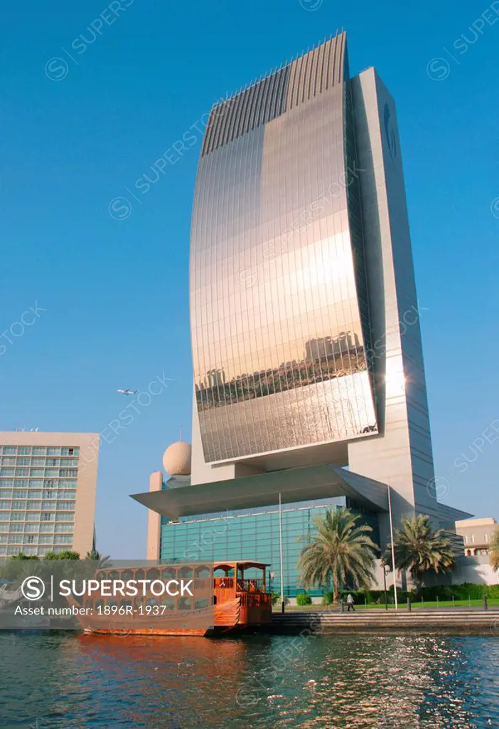 The National Bank of Dubai building with a wooden restaurant dhow moored in front of it, City of Dubai, Emirate of Dubai, United Arab Emirates.