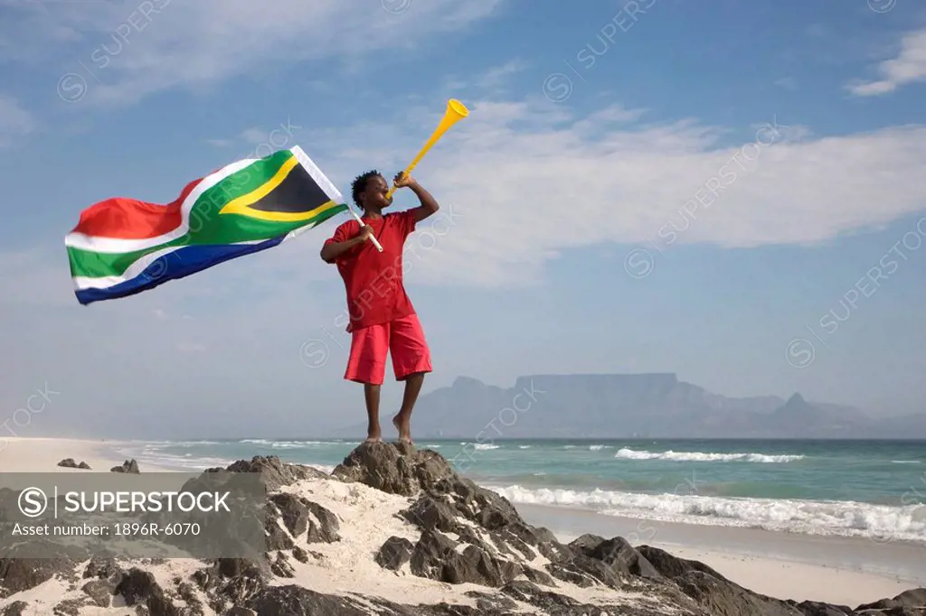 Young boy blowing on vuvuzela, while holding South African flag on beach, Table Mountain in background, Blouberg Beach, Cape Town, Western Cape Provin...