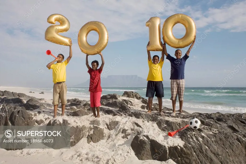 Boys standing on rocky outcrop, holding golden balloons shaped as numbers for 2010, Table Mountain in background, Blouberg Beach, Cape Town, Western C...