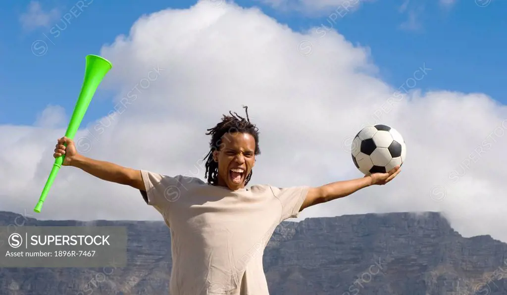 Young soccer fan cheering to camera with vuvuzela and soccer ball in each hand, Table Mountain in background, Table View, Cape Town, Western Cape Prov...