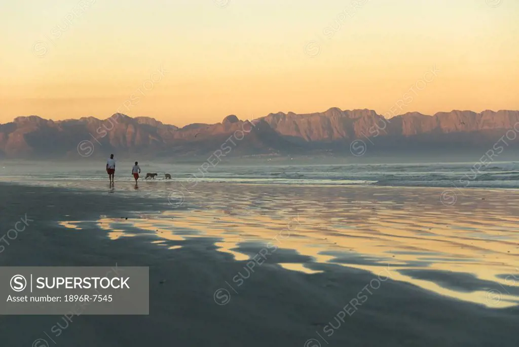 Two women walking dogs on beach, with Hottentot Holland Mountains in background at sunset, Muizenberg, Cape Peninsula, Western Cape Province, South Af...