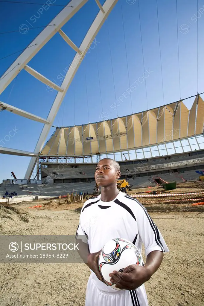 Football player holding ball in front of construction work behind him, Moses Mabhida Stadium, Durban, KwaZului_Natal Province, South Africa