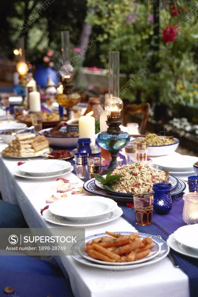 An outdoor dining table set for dinner with blue and white dinnerware, plates of food, and lit lanterns.