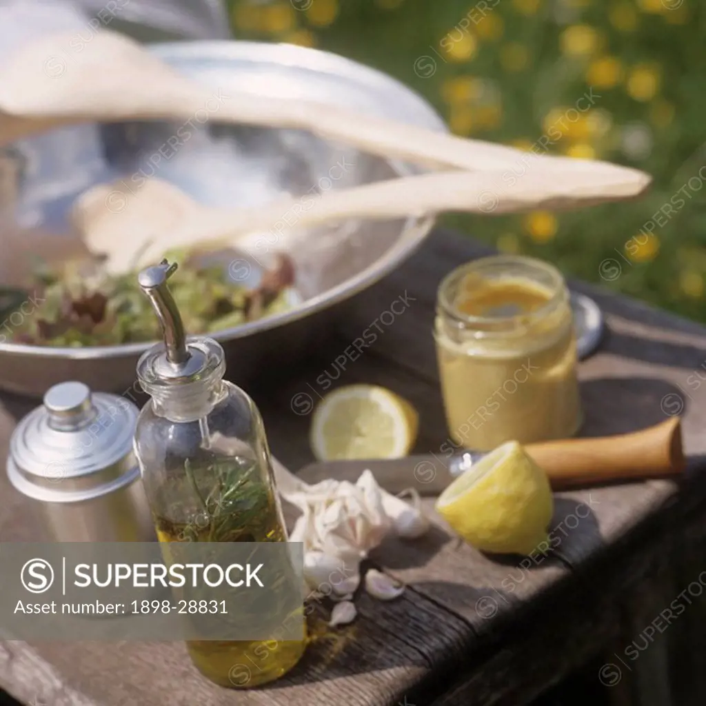 Bottle of oil with herbs, lemon, mustard, and salad in a bowl on a picnic table