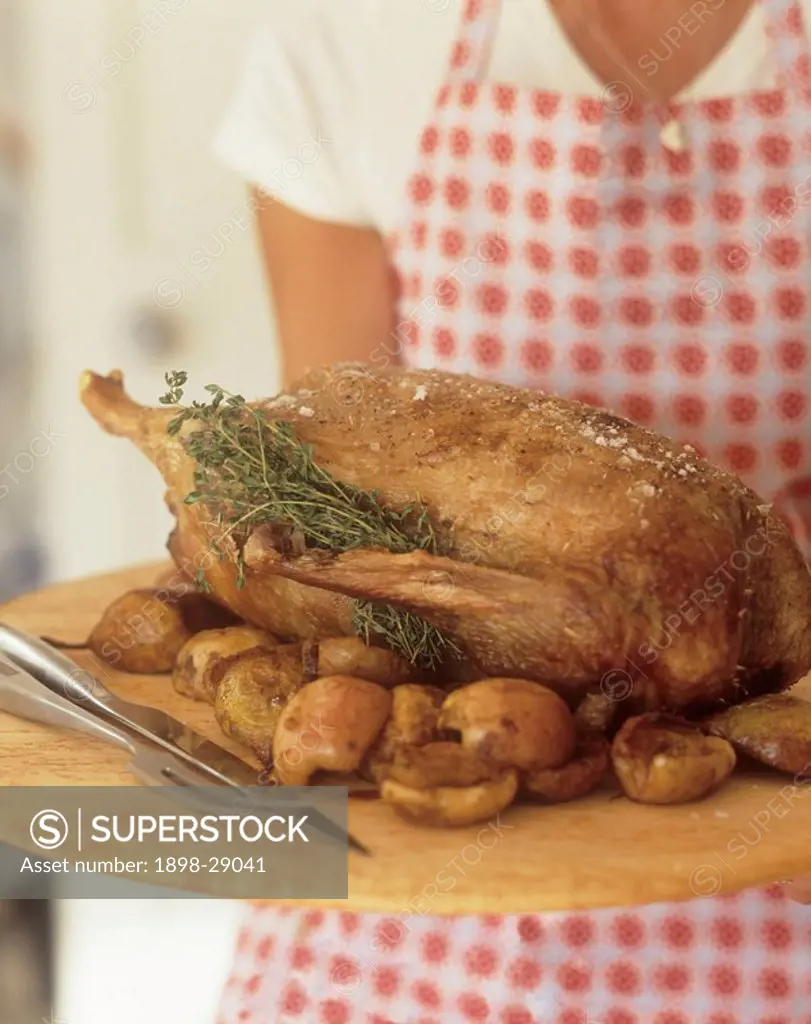 Woman holding roast duck with apple and herbs on wooden tray.