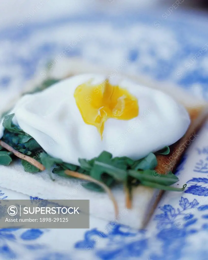 Close up of a poached egg on toast with green rocket leaves on a decorative blue china plate.