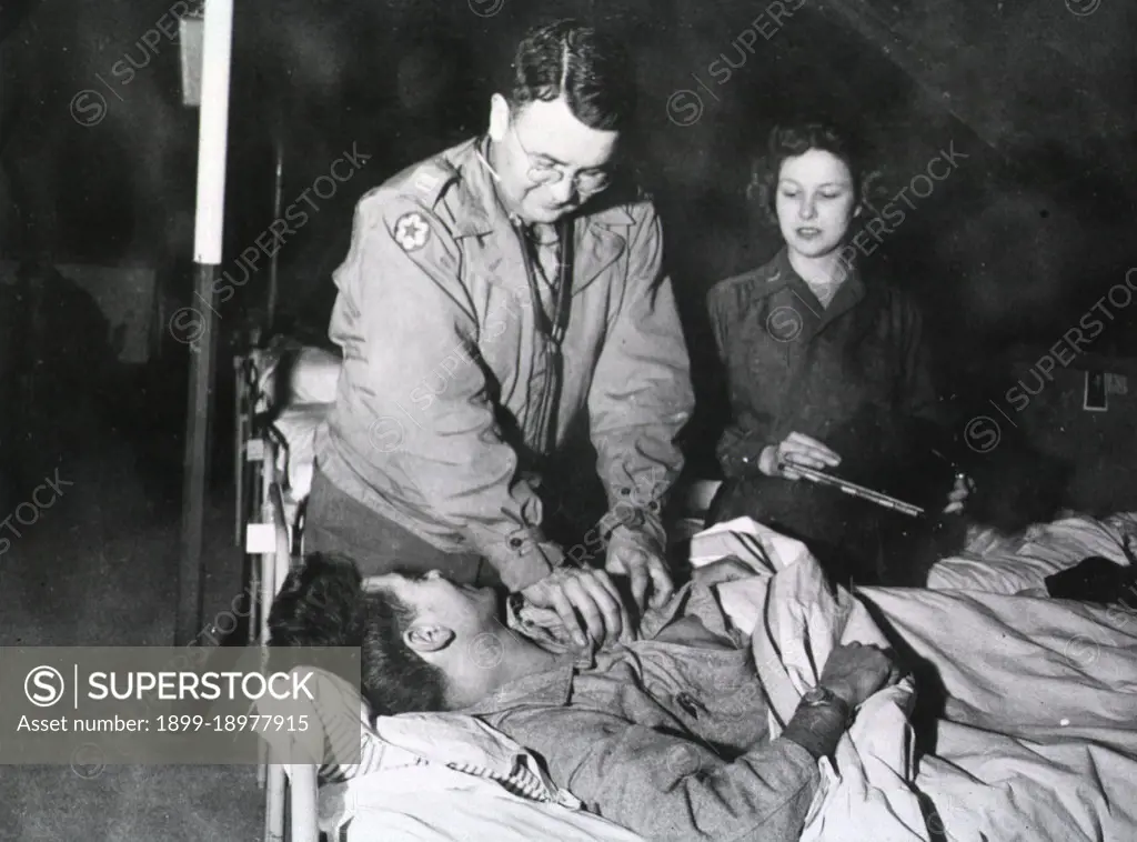 A doctor leans over and unbuttons the shirt of a wounded man lying in bed while a nurse holds a medical chart and watches the patient. 