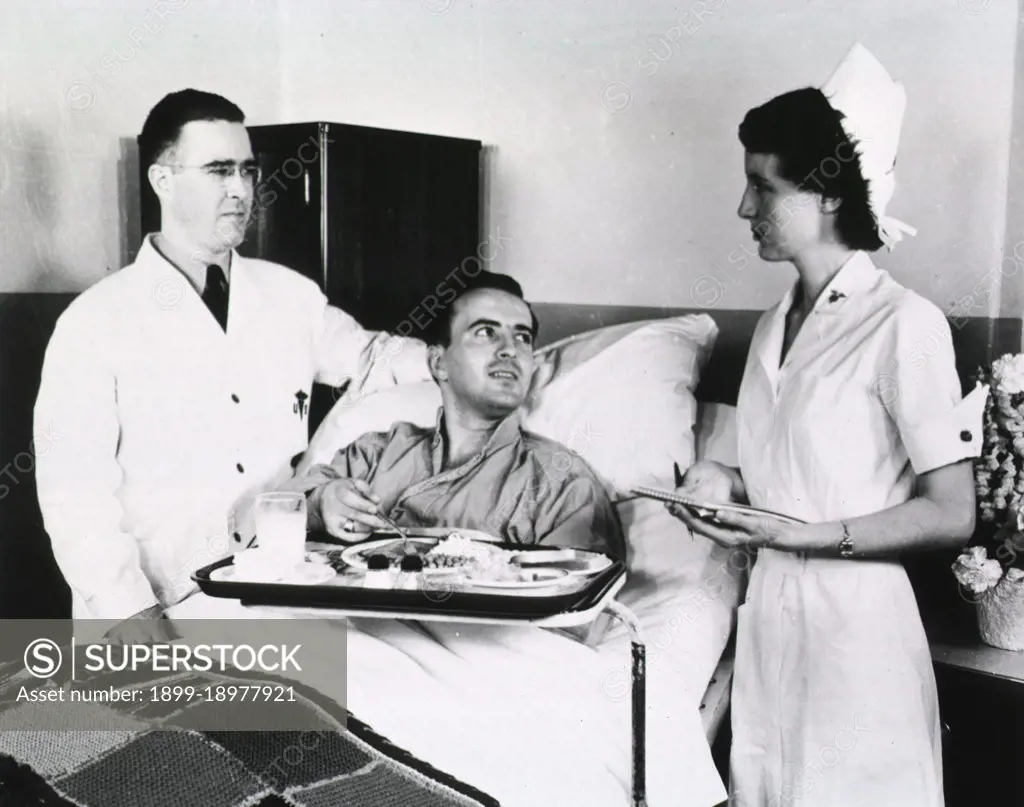 A male doctor and female nurse stand on either side of a hospital bed in which a male patient sits before a food tray. 