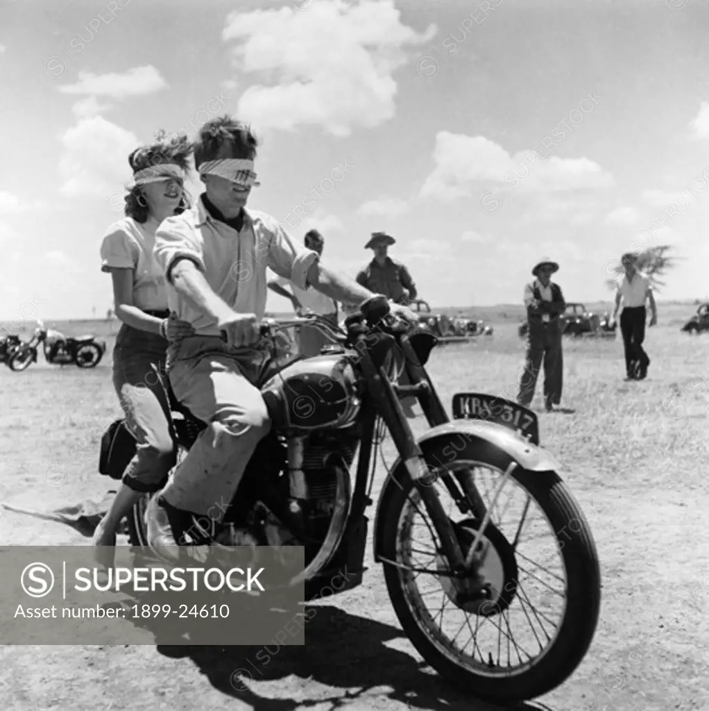 Blindfolded biking. A blindfolded couple sit astride a motorcycle at a motor sports gymkhana. Pat Dale sits in the driving seat with a female passenger riding pillion behind him. Kenya, 21 February 1954. Kenya, Eastern Africa, Africa.