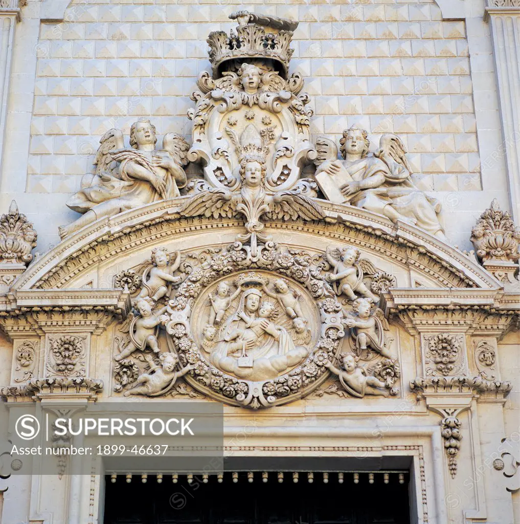 Convent of Santa Maria del Carmine, Lecce, by Manieri Mauro, Cino Giuseppe, 1711, 18th Century, Lecce stone. Italy: Puglia: Lecce: Lecce: Santa Maria del Carmine Convent. Detail. Exterior church Monastery of Santa Maria del Carmine facade superimposed orders Lower order doorway curved pediment carved tympanum/gable angels holding garlands garland flowers cherubs/putti angels plant frieze acanthus leaves cornice molding Madonna Virgin Mary Crowned Baby Jesus/Child Jesus/Christ Child crest/coat of