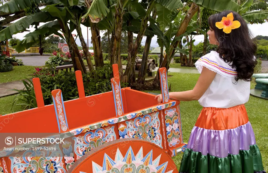 Costa Rican Woman In Traditional Dress Next To Traditional Carriage In And Around Costa Rica.