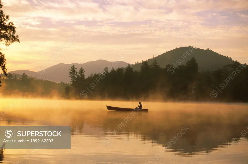 A Man In A Canoe Crosses A Misty Lake In A Mountainous Region