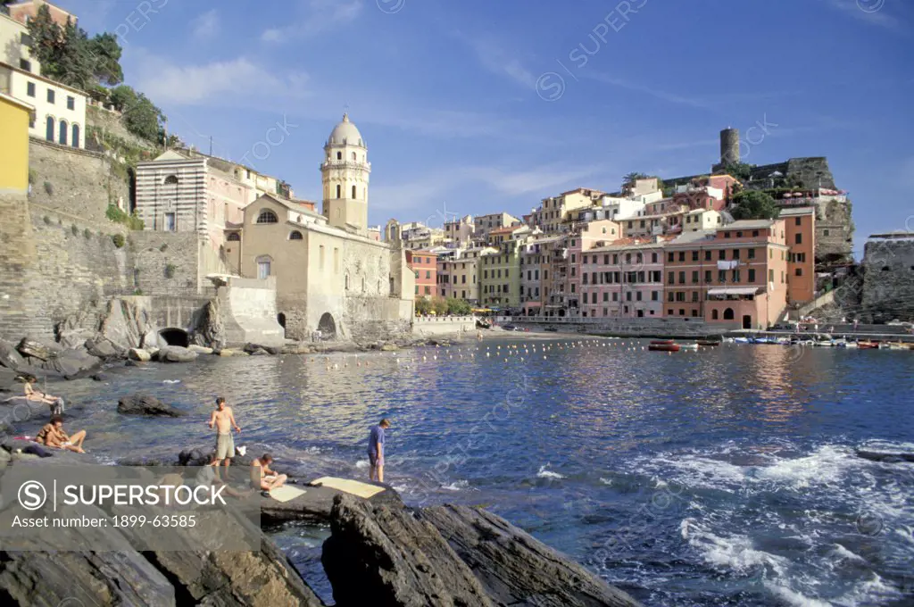Italy, Liguria, Cinque Terre, Vernazza. People Sitting On Rocks Along Water, Town Beyond
