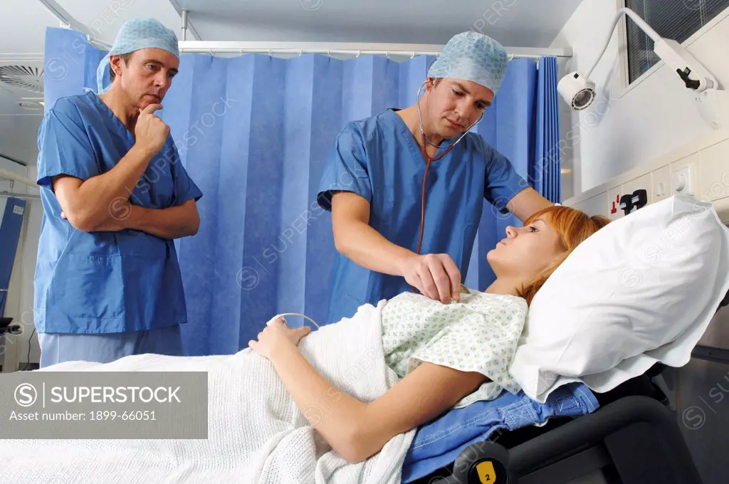 Two doctors, wearing blue surgical gowns and caps, using stethoscope to check breathing of young woman before operation