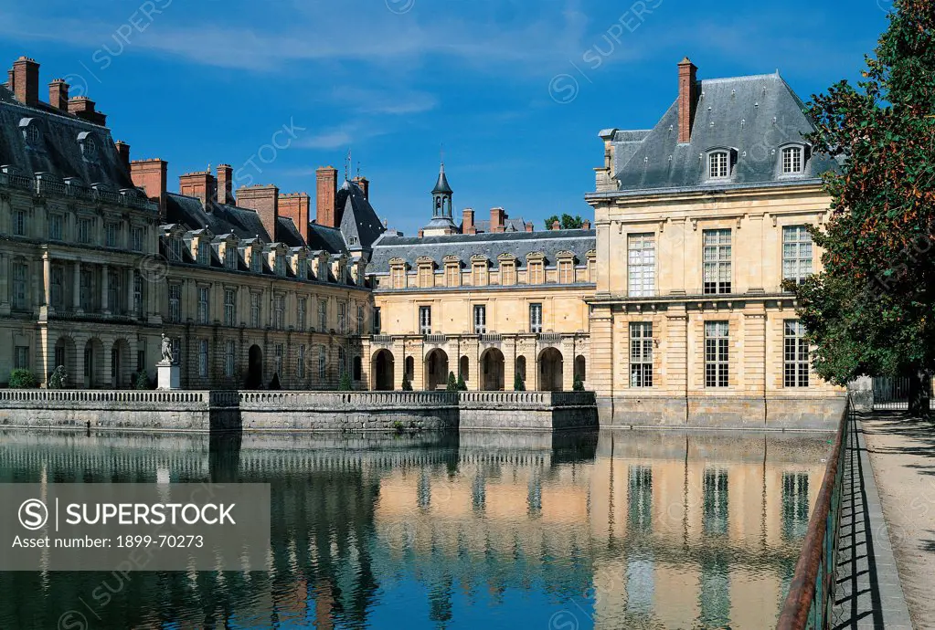 France, Fontainebleau, Palace of Fontainebleau. Detail. Exterior detail of Fontainebleau 'cour de la Fontaine' court, representing the facade and the wings where are tympanum windows. In the foreground, a pond with water and a fountain on the left. On the roof of the palace are slopes and chimneypots.