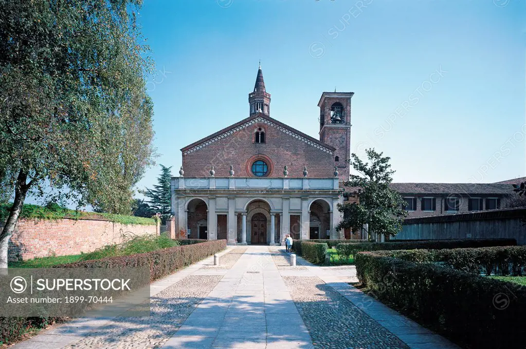 Italy, Lombardy, Milan. Whole artwork view. Frontal view of the approach alley and the facade of this Cistercian abbey that presents a bell tower, a portico, rose window and columns.
