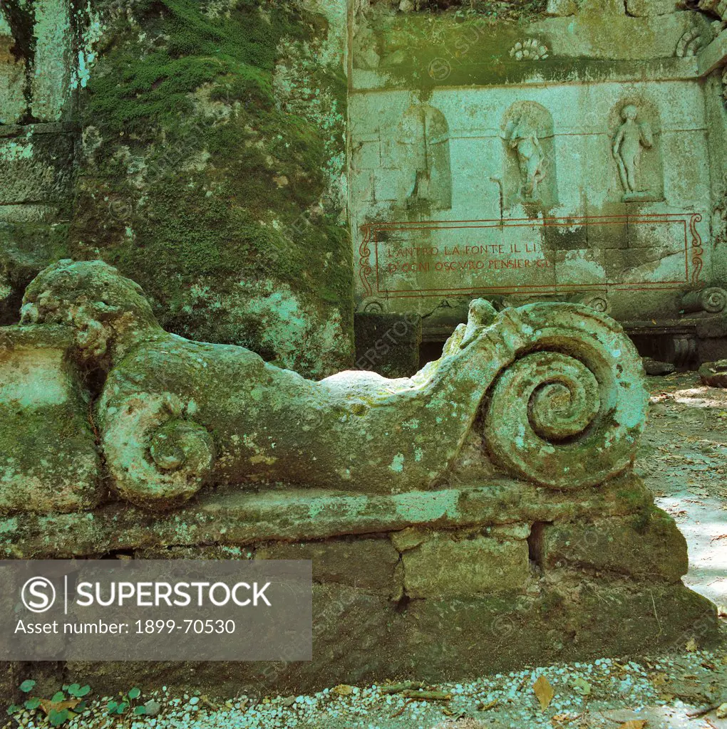Italy, Lazio, Bomarzo (Viterbo), Palazzo Orsini. Whole artwork view. View of a sculpted decoration covered with moss consisting of a human figure with a column and volutes. In the background there are three high-relief statues inside three niches.