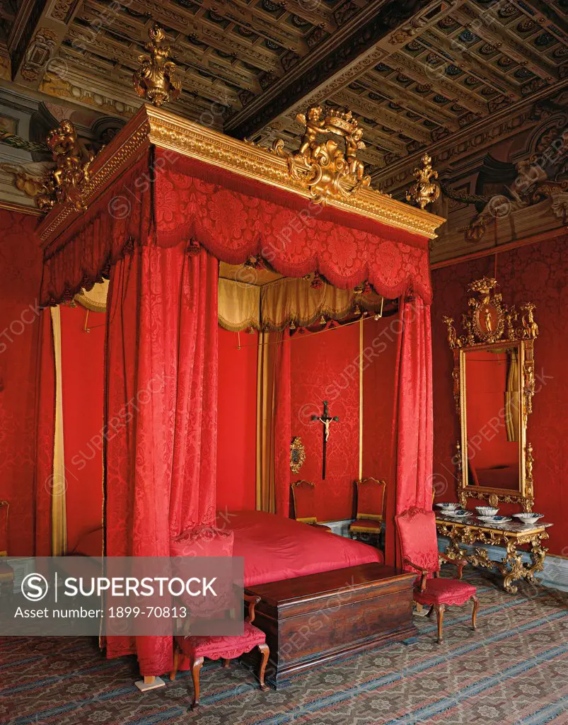 Italy, Tuscany, Pistoia, Palazzo Rospigliosi. Whole artwork view. A red damask bedroom with a foreshortening view of the four poster bed; on the carved and gilded wooden frame of the canopy stand two lateral vases and a central crown held by two cherubs.