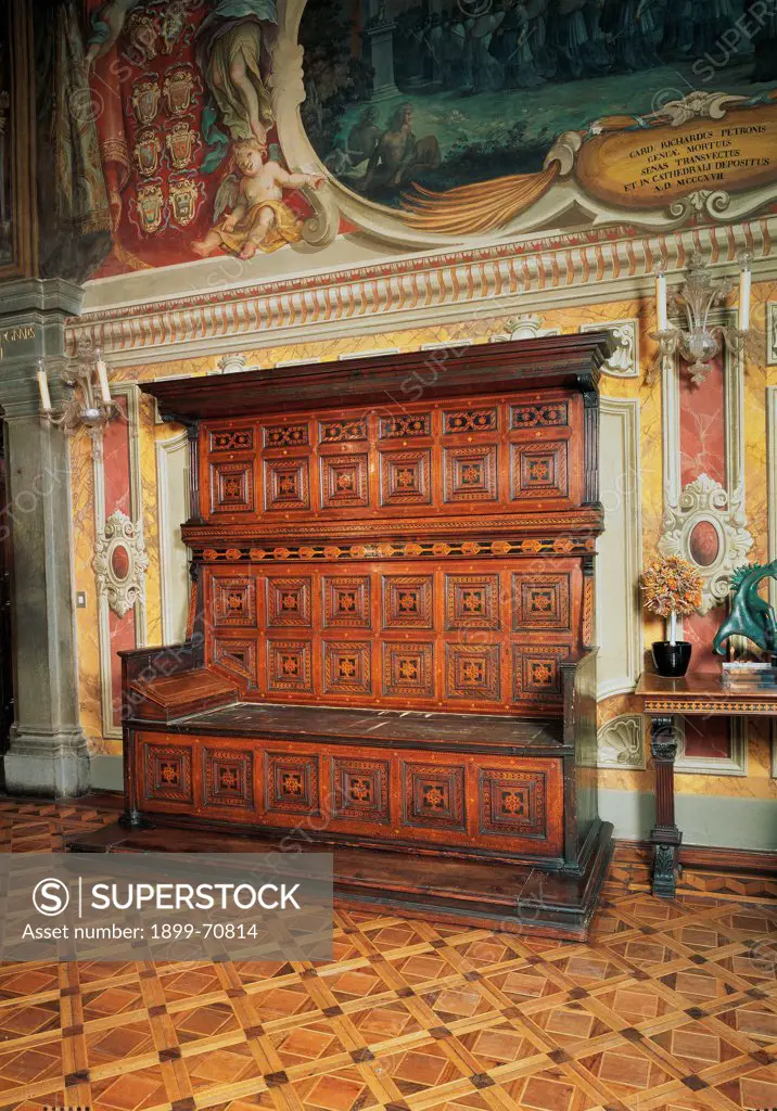 Italy, Tuscany, Siena, Palazzo Pubblico. Whole artwork view. View of a throne-type settle bed with squared inlaid decorations on the surface; it's located in a room with painted walls and a parquet floor.