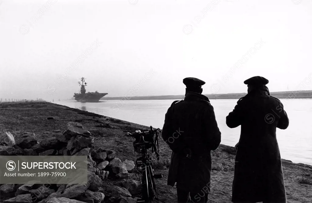 Two military standing along the Suez Canal, watching a U.S. aircraft carrier arrives in the distance. Arab-Israeli Six-Day War. Egypt, 1967.
