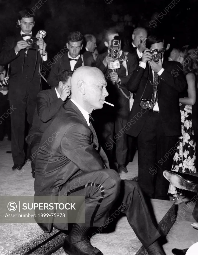 American actor Yul Brynner (Yuliy Borisovich Bryner) sitting on a step and smoking a cigarette photographed by some photographers. Taormina, 1966.