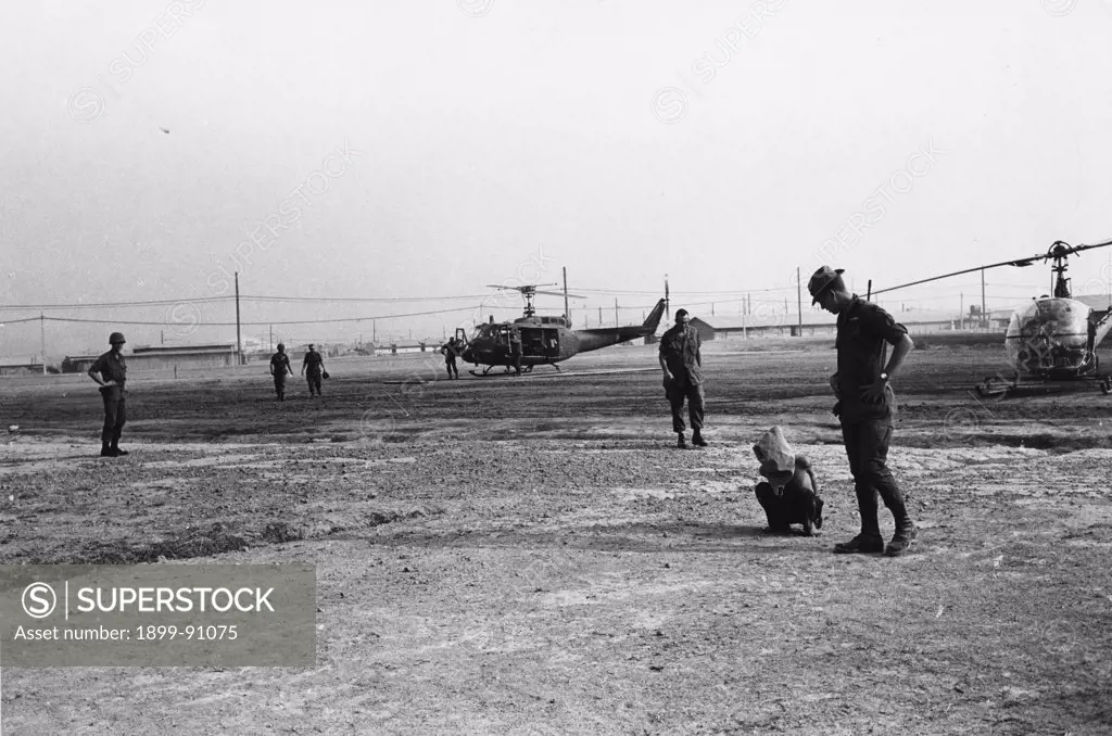 Viet Cong prisoner, tied and gagged (infringement of the Geneva Convention), in an American camp. In the background some helicopters and soldiers. Vietnam, 1968.