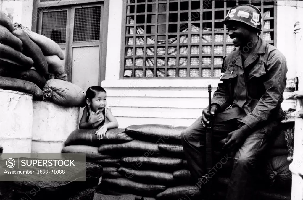 An American sentry is smiling as he seats guard in a street of the city, holding a gun in his hand. Next to him, a curious young girl who is leaning against the sand sacks. War in Vietnam (1960-1975). Saigon (Vietnam), 1968.