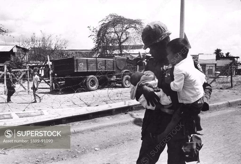 An American soldier is keeping a young boy and a newborn in his arms. In the background, a truck and other two children behind a barbed-wire fence. War in Vietnam (1960-1975). Saigon (Vietnam), 1968.