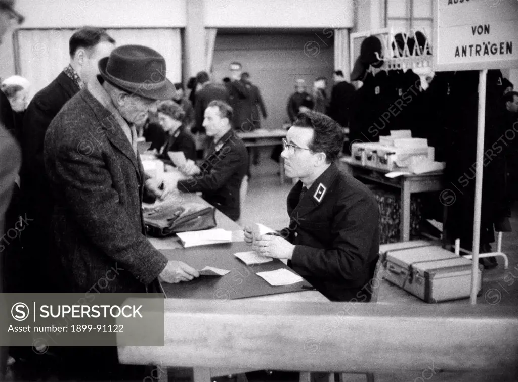 An old German man, wearing a coat and a hat, is showing some papers to the policeman seated at the desk; the man is asking the pass to go to East Berlin for Christmas time. West Berlin (Germany), December, 1963.