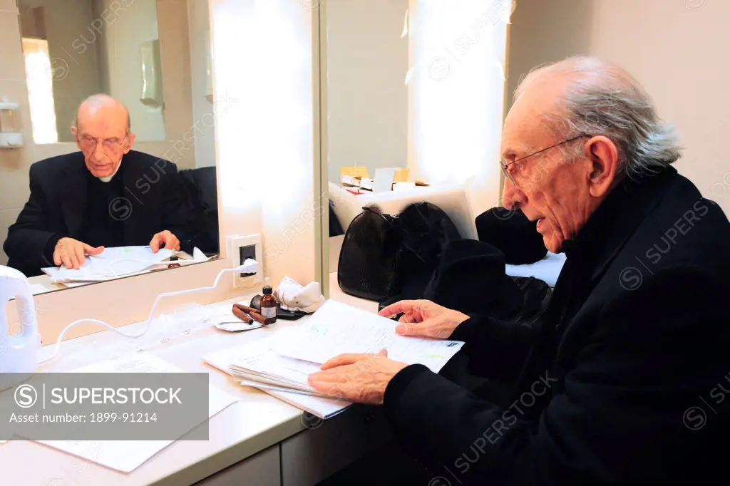 Don Andrea Gallo seated in front of a mirror of a dressing room while he is turning over the pages of a block of paper. Italia, 2012.