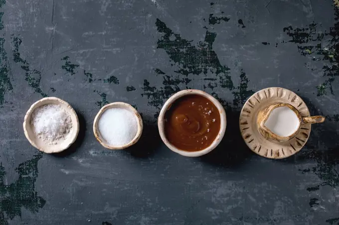 Homemade salted caramel sauce with grains of fleur de sel salt. Ingredients in ceramic bowls in row. Grey textured canvas background. Flat lay. space