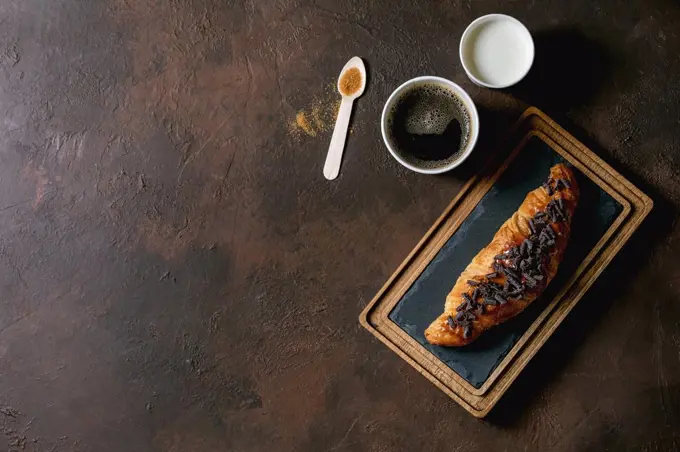 Traditional french puff pastry and chocolate. croissant on slate wooden board with paper cups of coffee and milk. recycled wooden spoon of cane sugar over dark texture background. Flat lay. space