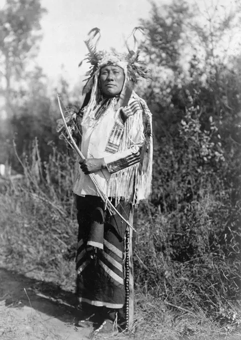 Edward S. Curtis Native American Indians -  Long Time Dog, full-length portrait, wearing headress, buckskin shirt, and leggings with brass bells ca. 1908. 