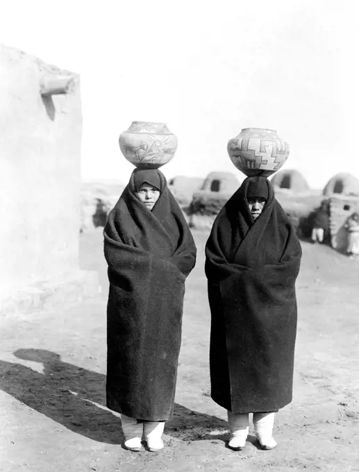 Two females, full length, wearing blankets, pots on heads. 