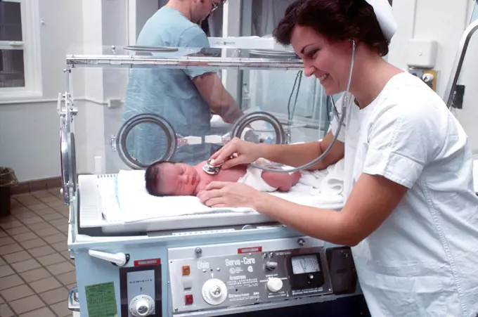 1977 - A nurse examines an infant in an incubator. 